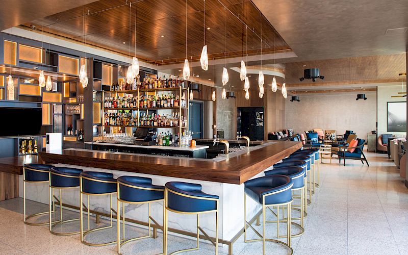 A modern bar with blue barstools, a well-stocked bar, hanging lights, and a seating area in the background with a mix of chairs and couches.