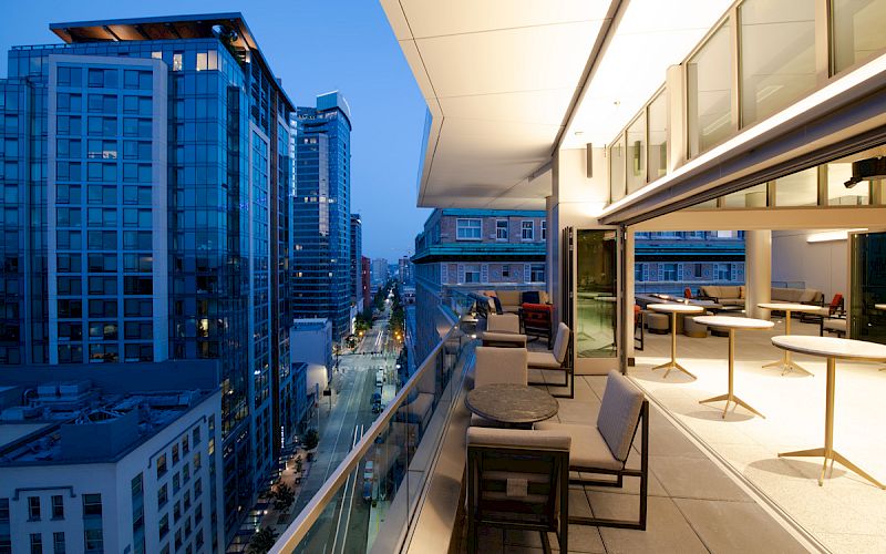 The image shows a modern urban scene with tall glass buildings and a furnished balcony overlooking the street below during the evening.