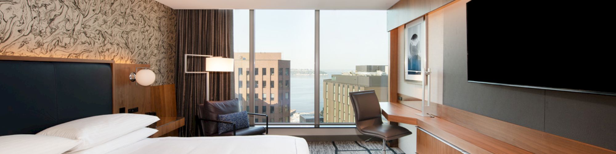 This image shows a modern hotel room with a large bed, a wall-mounted TV, a desk with a chair, and a window with a cityscape view.
